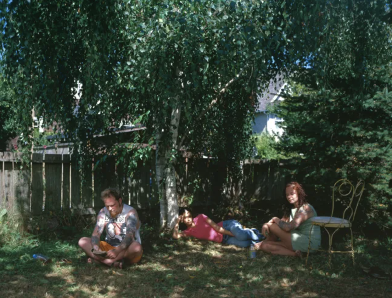 Drei Personen sitzen im Schatten eines großen Baums im Garten.